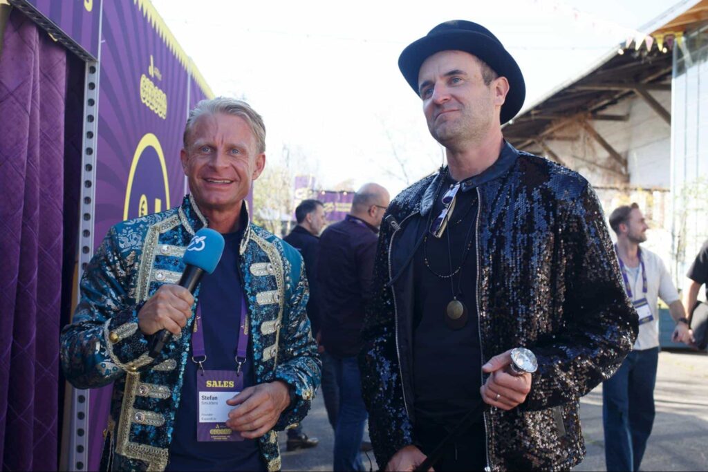 Two men in colorful, festive outfits at an outdoor event, one holding a microphone